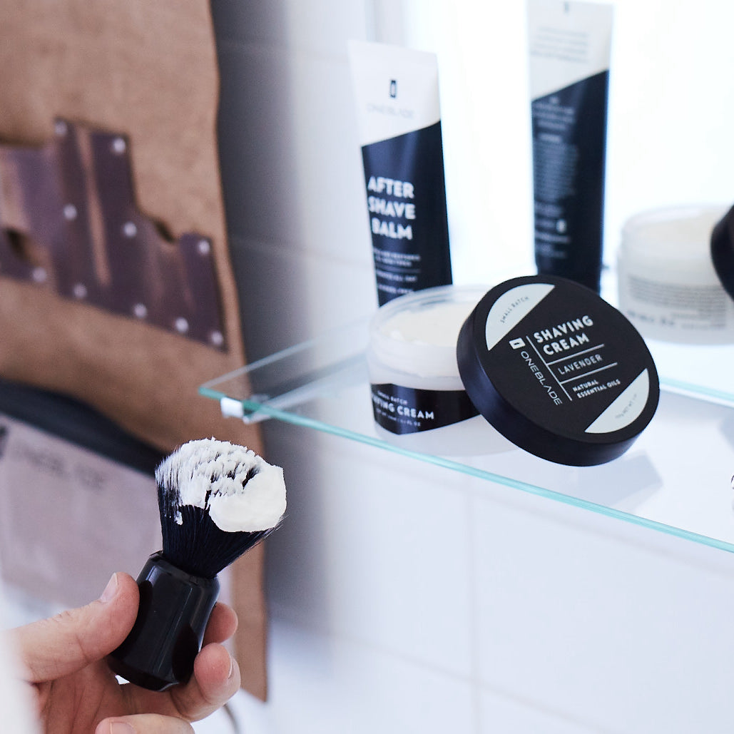 hand holding a shaving cream brush, with after shave balm on a shelf near by