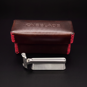 A silver metal safety razor with a black handle grip, displayed in front of its brown leather carrying case with red stitching. The embossed brand name 'ONEBLADE' is visible on the top of the case against a dark background.